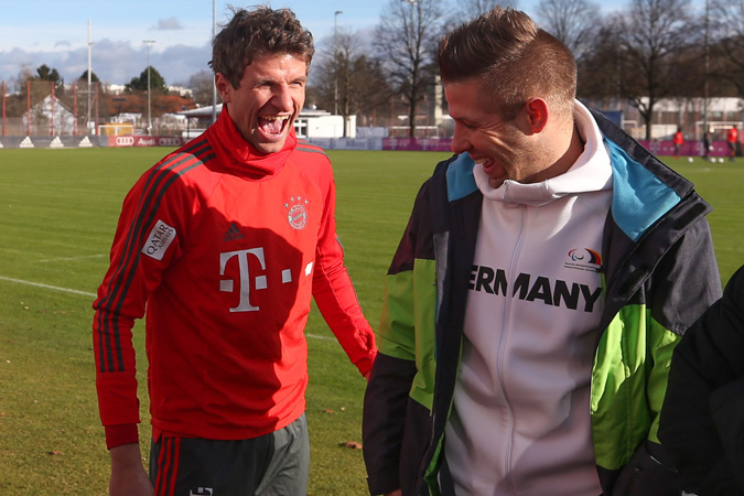 Fußball-Nationalspieler Thomas Müller und Amputierten-Fußball-Nationalspieler Christian Heintz lachend auf einem Rasenplatz